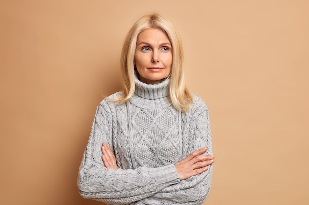 Photo of beautiful middle aged female with blonde hair keeps arms folded thinks about something contemplates over future plans wears warm sweater.