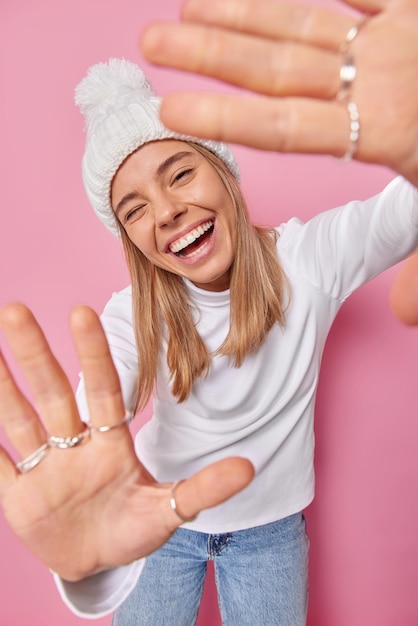 Free photo photo of carefree cheerful pretty teenage girl smiles happily winks eye at camera raises palms and has fun wears casual turtleneck jeans and hat poses against pink foolishes around