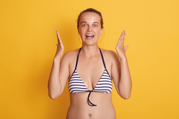 Photo of charming young woman in striped swimsuit and showing size with hands isolated over yellow wall, looks excited.