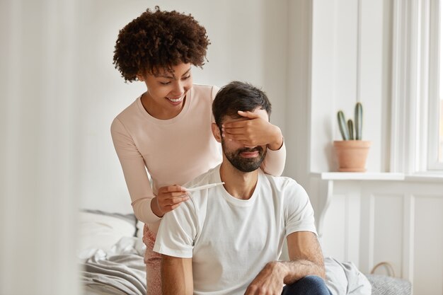 Photo of cheerful glad dark skinned young woman covers husbands eyes, wants to make unexpected surprise, shows positive pregnancy result, pose on bed, going to become parents. Maternity concept