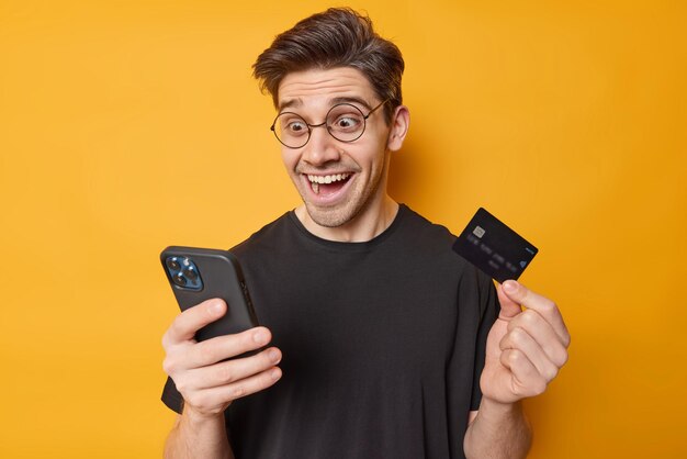 Photo of cheerful surprised adult man stares in smartphone with unbelievable gaze shocked to receive big amount of money wears spectaces and casual black t shirt isolated over yellow background