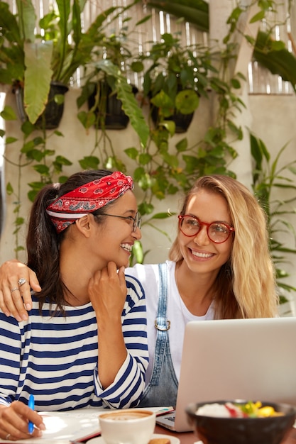 Photo of cheerful women prepare for examination session, do home assignment together, write in notebook, enjoy coffee