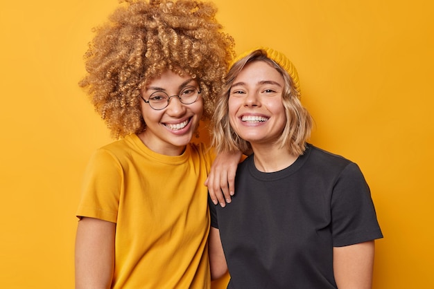 Photo of cheerful young beautiful women stand next to each other dressed in casual t shirts smile happily isolated over vivid yellow studio background People friendship positive emotions concept