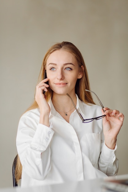 Photo of a cheerful young blonde business woman in office indoors work with laptop and mobile phone.