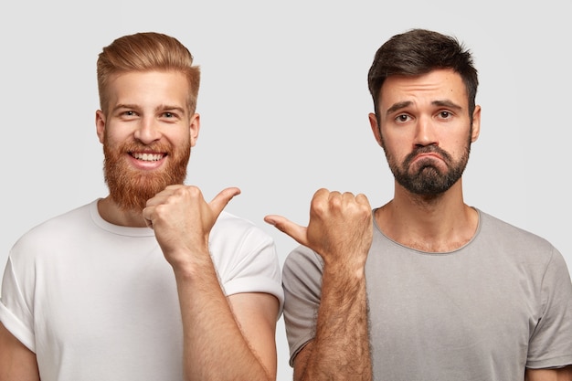 Free Photo photo of cheerful young ginger man and displeased unshaven caucasian guy point with thumbs at each other, work together as team, isolated over white wall. friendship and emotions concept