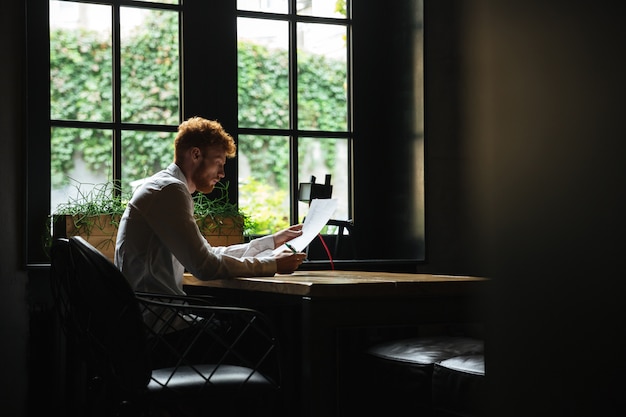 Photo of concentrated readhead bearded businessman reading contract, sitting at the cafeteria