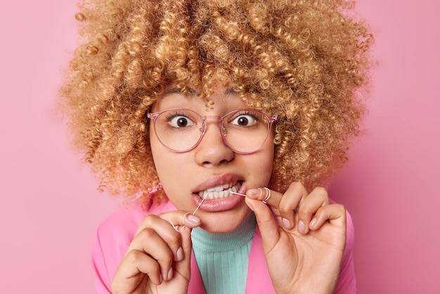 Free Photo photo of curly haired woman cleans teeth with dental floss takes care of oral hygiene wears transparent glasses dressed in formal clothes isolated over pink background dental health concept