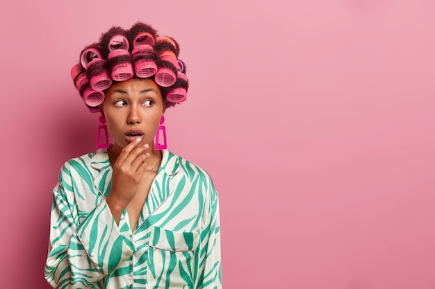 Photo of dark skinned woman gets hair curled, wears curlers and makes hairstyle at home, keeps hand on opened mouth, wears casual clothing, poses against pink wall, blank empty space aside