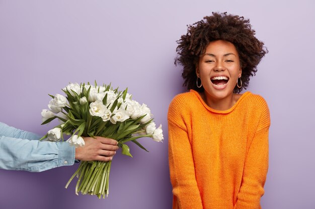 Photo of delighted African American lady laughs sincerely, gets flowers from husband or boyfriend