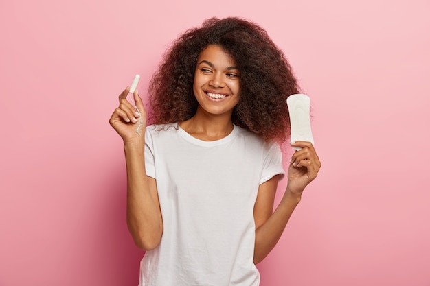 Photo of delighted African American woman holds tampon and sanitary napkin, dressed in white t shirt, isolated over pink wall. Women, pms