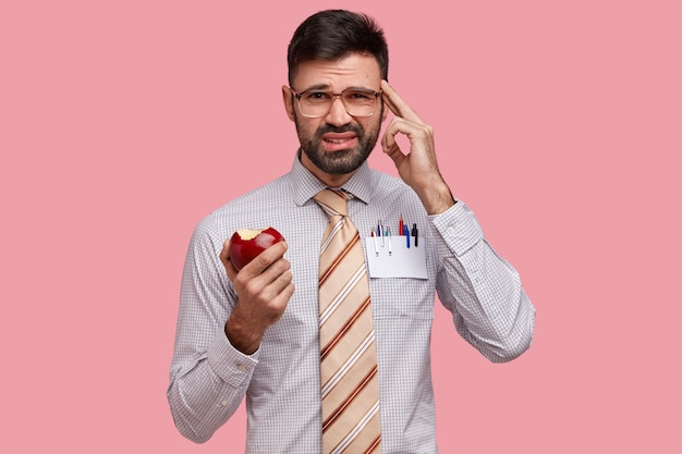 Photo of discontent young Caucasian man keeps finger on temple, dressed formally, eats apple, recollects something in mind