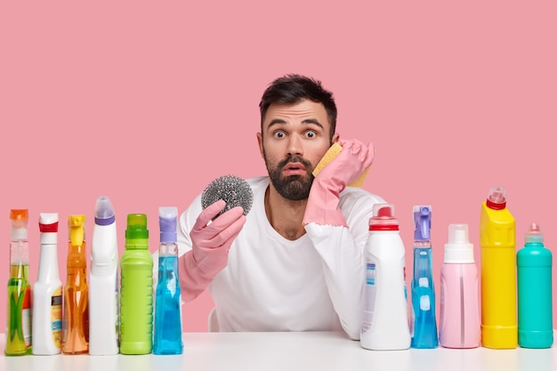 Photo of displeased bearded man in rubber protective gloves holds rag for cleaning kitchen stove, looks embarrassed