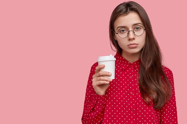 Photo of displeased sad woman drinks coffee, has sleepy expression, enjoys fresh drink