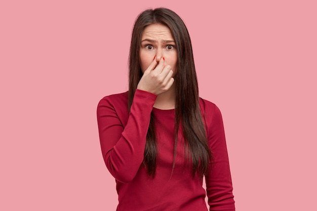 Photo of displeased woman closes nose with reek, feels terrible smell from garbage, wears red clothes