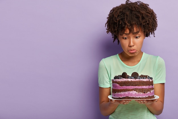 Photo of dissatisfied Afro American woman holds plate of blueberry sweet cake, purses lower lip, has no good will, wants to eat delicious dessert but keeps to diet