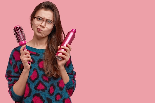 Photo of dissatisfied woman frowns face, feels displeased, carries comb and hairspray for making haircut, dressed in domestic clothes