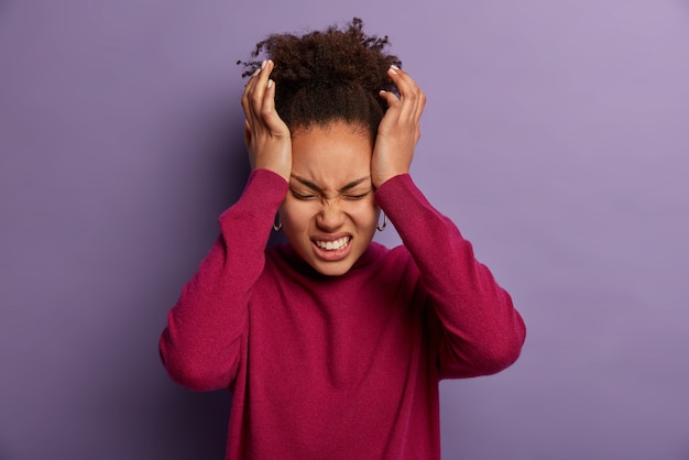 Photo of dissatisfied woman keeps hands on temple, suffers from unbearable headache, clenches teeth with pain, being exhausted after work, wears burgundy turtleneck, isolated on purple wall.