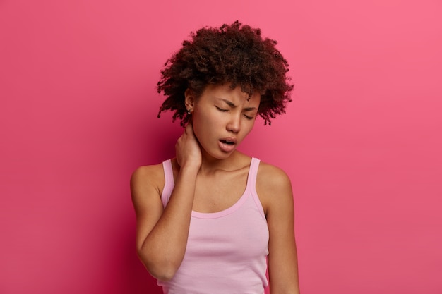 Photo of distressed woman touches neck, suffers from back or spine disease, yawns and closes eyes, has tired expression, dressed casually, has painful feelings, isolated over pink wall