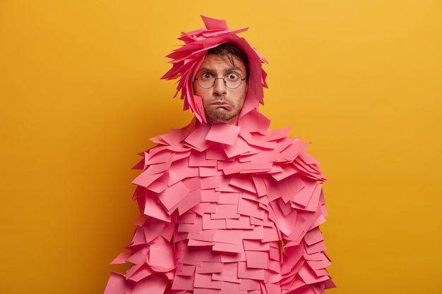 Free photo photo of embarrassed nervous male advertiser bites lips, feels dissatisfied and tired, covered with pink sticky notes over body, poses indoor against yellow wall, has puzzled expression