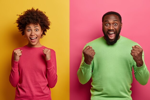 Free photo photo of emotional dark skinned female and male clench fists, exclaim and support favorite football team, have overjoyed face expressions, dressed in casual wear, isolated on yellow and pink wall