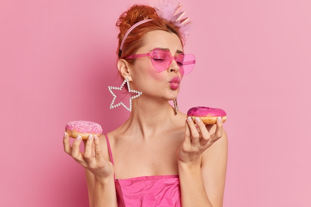 Photo of fashionable redhead glamour European woman keeps lips folded holds two appetizing doughnuts wants to eat sweet dessert