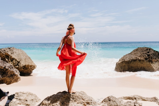 Photo from back of shapely tanned girl standing on big stone. Outdoor shot of graceful female model playing with her red dress and looking at ocean waves.