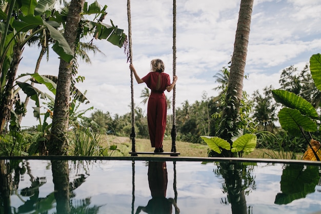 Photo from back of slim woman in long dress looking at rainy sky. Outdoor shot of shapely female model enjoying nature views at resort.