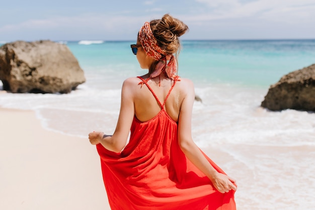 Photo from back of tanned gorgeous woman looking at sea. Outdoor portrait of amazing caucasian girl in red summer dress standing.