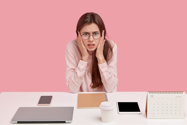 Photo of frustrated Caucasian woman dressed in elegant shirt, poses at workplace alone, has neatly arranged things, wears round spectacles