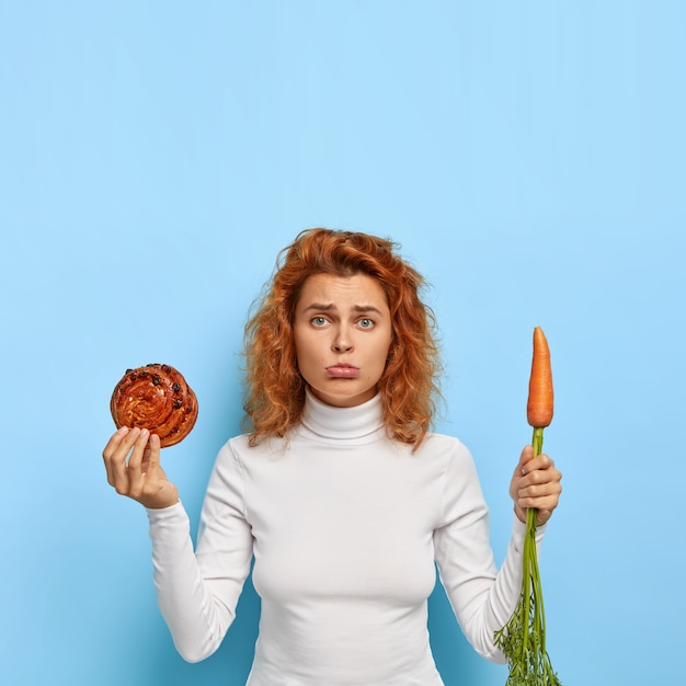 Free Photo photo of frustrated disappointed young woman keeps to diet, purses lower lip, makes hard choice between bun and carrot, healthy nutrition and junk food, has curly red hair, appealing appearance