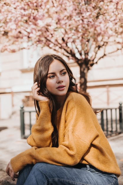 Free photo photo of girl in yellow sweater on background of sakura. woman in jeans posing outside. snapshot of lady in stylish outfit enjoying spring weather