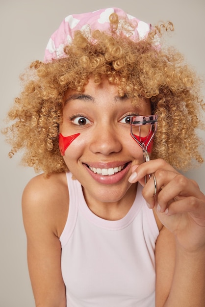 Photo of glad curly haired woman uses eyelashes curler wears bathhat and casual t shirt applies red beauty patches under eyes undergoes skin care treatment prepares for date isolated over grey wall