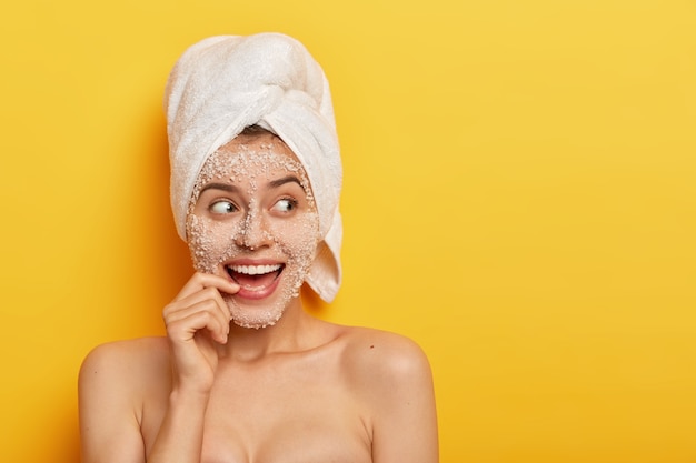 Free Photo photo of glad european girl with toothy smile, uses sea salt for spa procedures, takes shower, has smooth healthy skin, looks away, wears white towel, isolated over yellow background. beauty concept