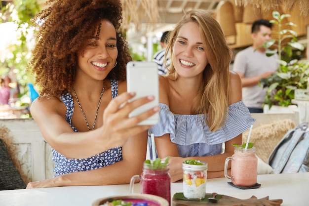 Photo of glad mixed race females have interracial friendship, pose at camera of modern cell phone, make selfie while have rest in cozy terrace bar, enjoy fresh drinks. People, ethnicity and leisure