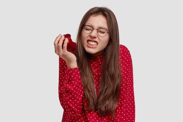 Photo of good looking woman clenches teeth, bites fresh red apple, dressed in elegant shirt, wears spectacles