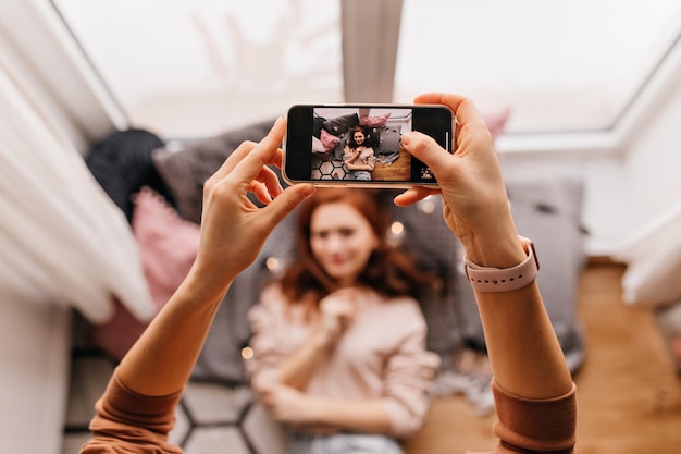 Free photo photo of hands holding smartphone and making photos. indoor portrait of red-haired lady posing for her friend.