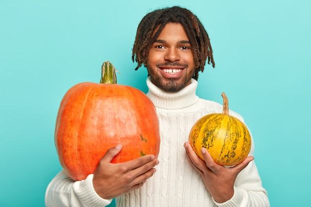 Photo of handsome cheerful hipster man with dreadlocks, toothy smile, holds two pumpkins of different size, wears warm white sweater, isolated over blue wall