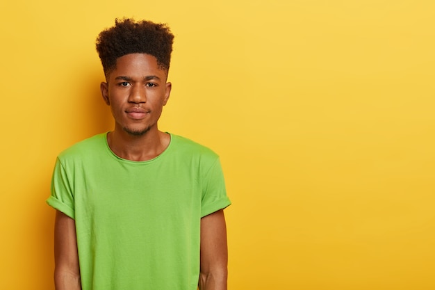 Photo of handsome teenage boy with dark skin, curly hairstyle, wears casual green t shirt, looks with calm serious expression