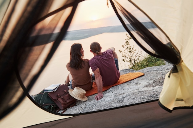 Photo of happy couple sitting in tent with a view of lake during hiking trip.