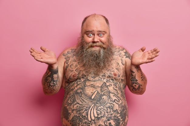 Free photo photo of hesitant cheerful man spreads palms sideways, says i dont know, being happy and confused, has big tummy, tattooed body, doesnt know how to be fit and loose weight, isolated on pink wall