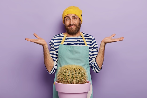 Free Photo photo of hesitant male florist spreads palms sideways, has unaware face expression, wears special uniform, isolated over purple background. botany concept