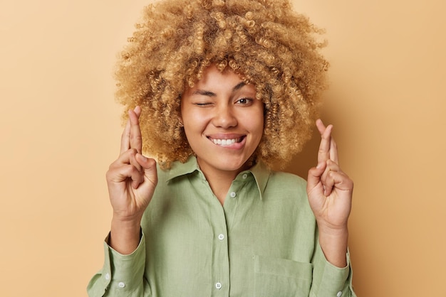 Free Photo photo of hopeful lwoman bites lips keeps fingers crossed believes in good luck makes cherished wish prays for miracle hopes for good things to happen wears green linen shirt isolated on beige wall