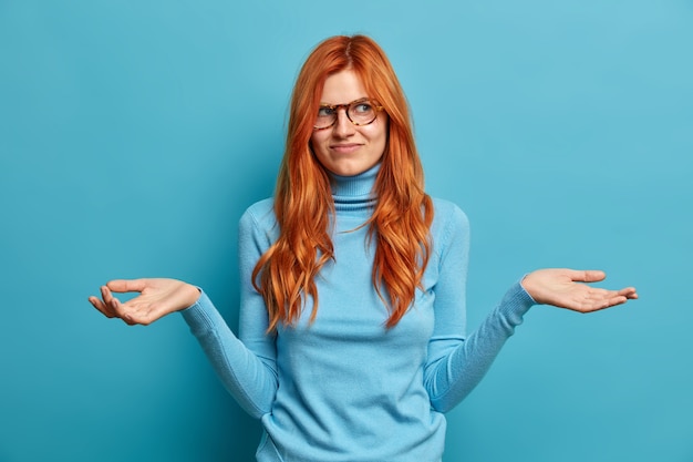 Free Photo photo of indifferent good looking woman with long red hair spreads palms and looks clueless cannot decide what to do wears casual clothes.