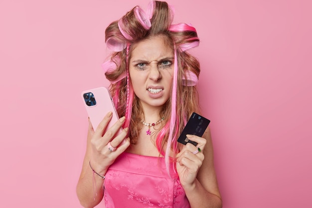 Photo of irritated young woman frowns face clenches teeth applies hair rollers holds mobile phone and credit card pays online wears dress has annoyed expression isolated over pink background