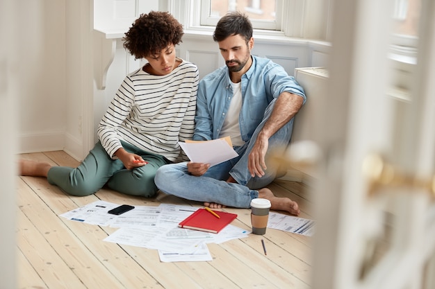 Free Photo photo of mixed race coworkers study financial bills, wear comfortable clothes, pose on floor, discuss future work and collaboration, surrounded with documents, coffee and cellular. working concept