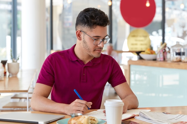 Photo of mixed race male student writes necessary information in notepad from daily newspaper, creates simmilar article, sits indoor against cafe interior, drinks take out coffee, learns indoor