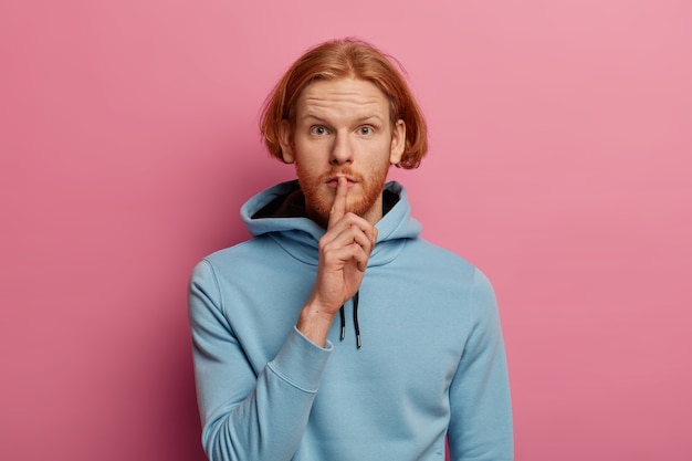 Photo of mysterious serious looking man with ginger hair and beard makes silence gesture, asks not tell his secret information, presses index finger to lips, wears blue hoodie looks directly 