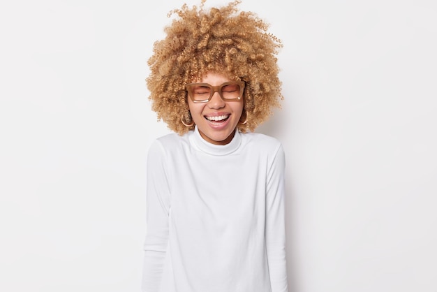 Free photo photo of overjoyed curly haired european woman laughs out smiles toothily keeps eyes closed wears spectacles and turtleneck isolated over white background. happy emotions and feelings concept