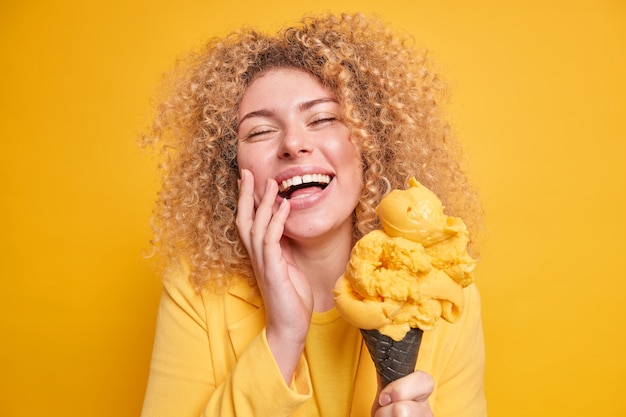 Free photo photo of overjoyed curly haired woman feels amused smiles broadly keeps eyes closed enjoys eating delicious ice cream of mango flavor poses with yummy frozen dessert isolated on yellow wall.