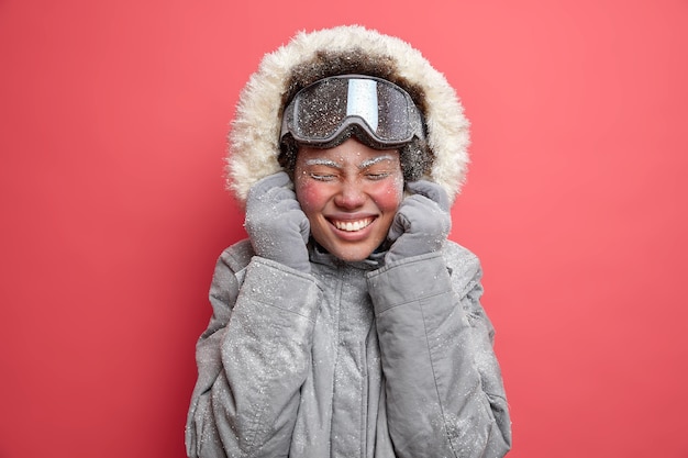 Free Photo photo of overjoyed woman wears hood of grey jacket smiles pleasantly has red face covered with frost goes skiing during december.